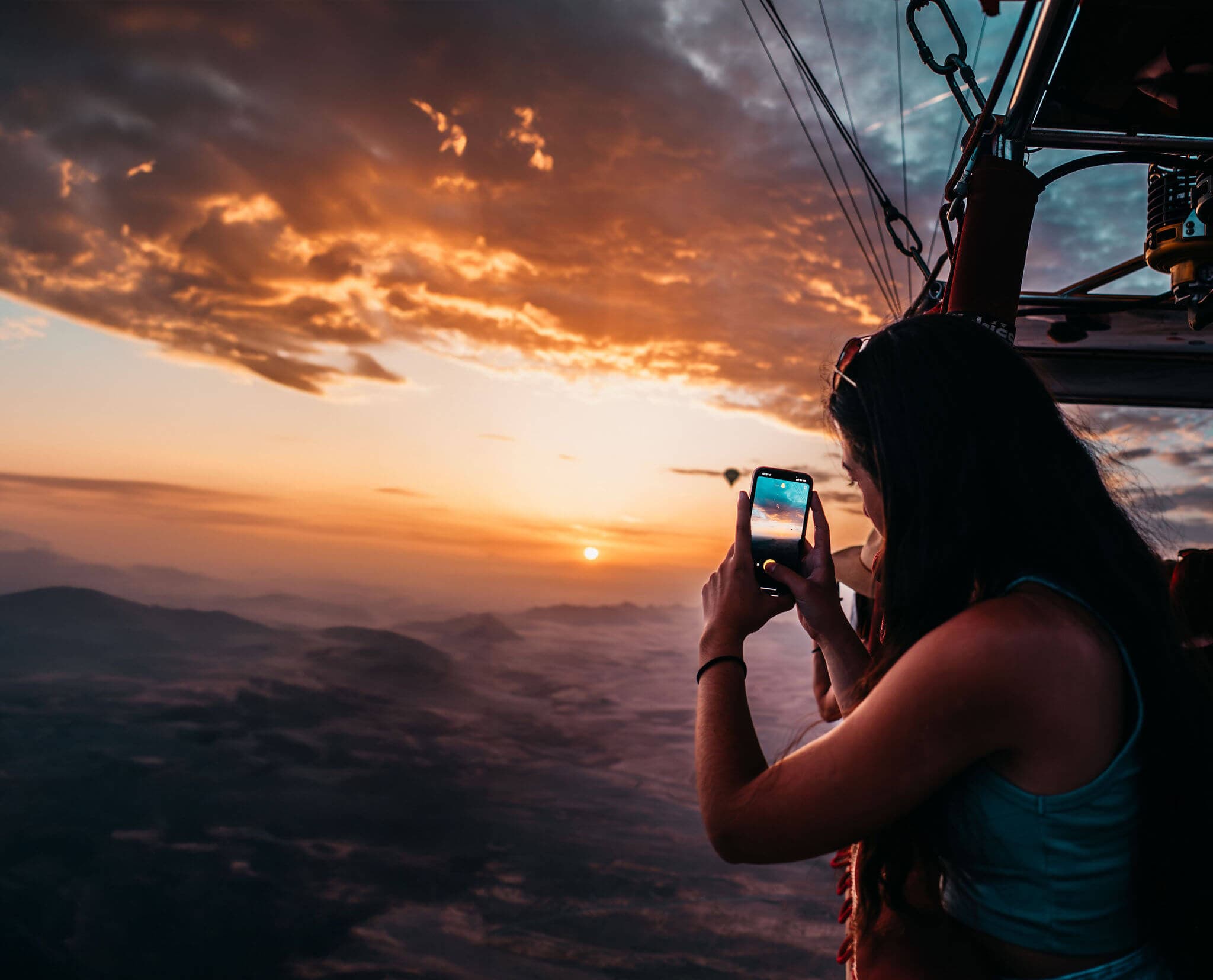 A traveller photographing the sunrise on her phone from a hot air balloon, high above the hazy Marrakech plain, with a second balloon in the distance