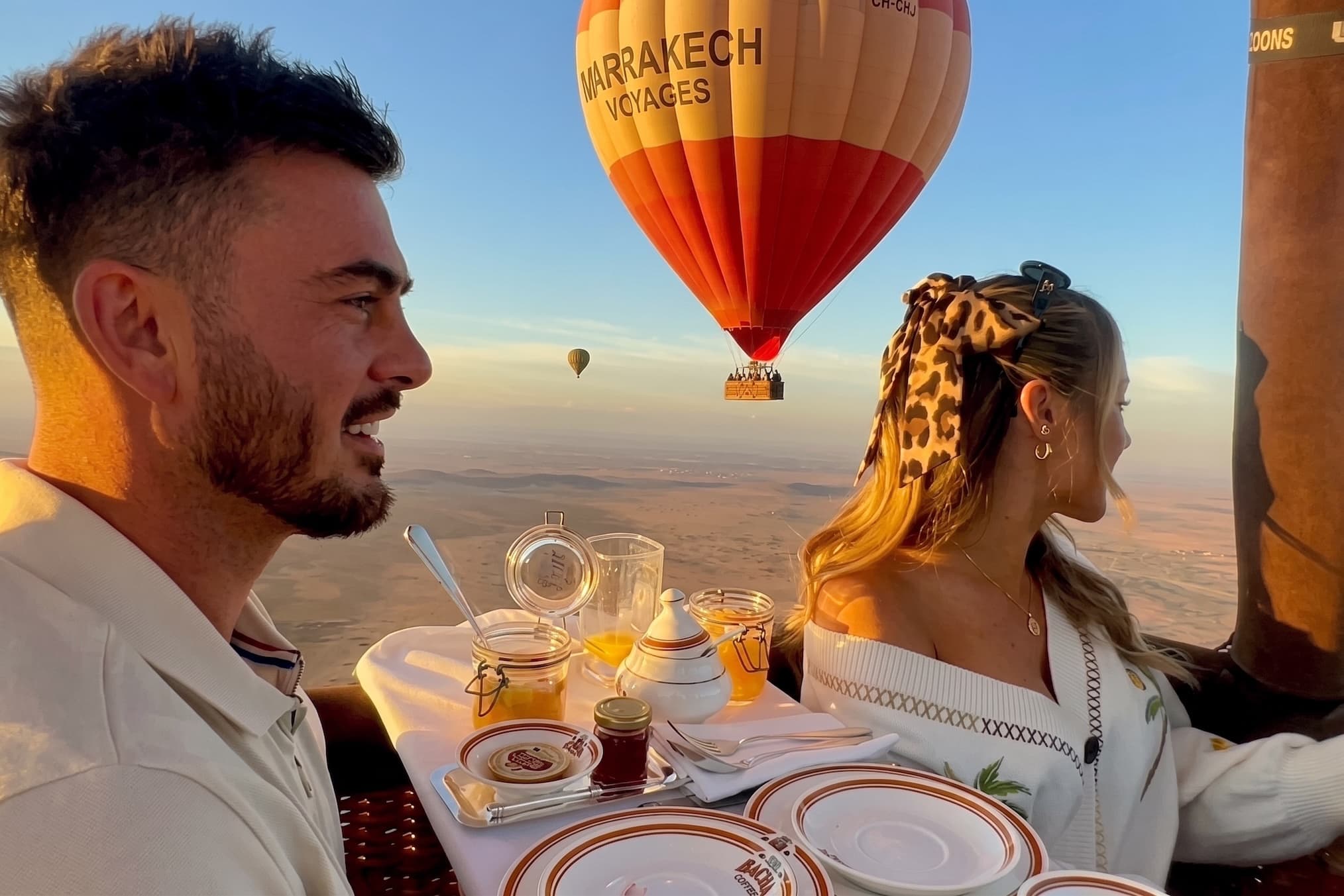 A couple at a laid breakfast table inside the balloon basket, another balloon in the sky behind